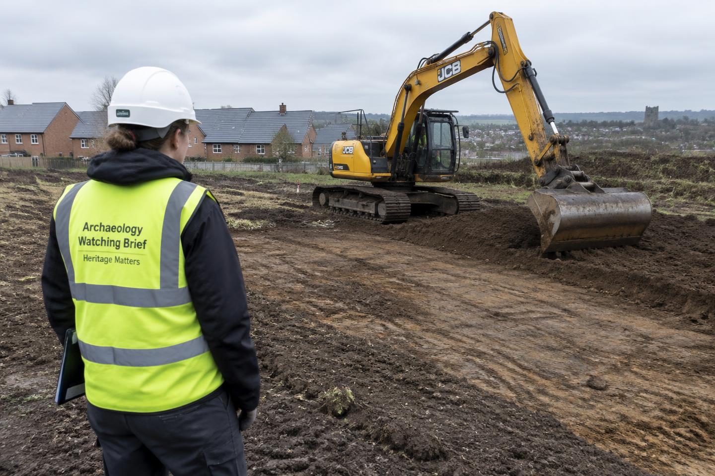 Archaeologist in hi-vis monitoring an excavator stripping topsoil